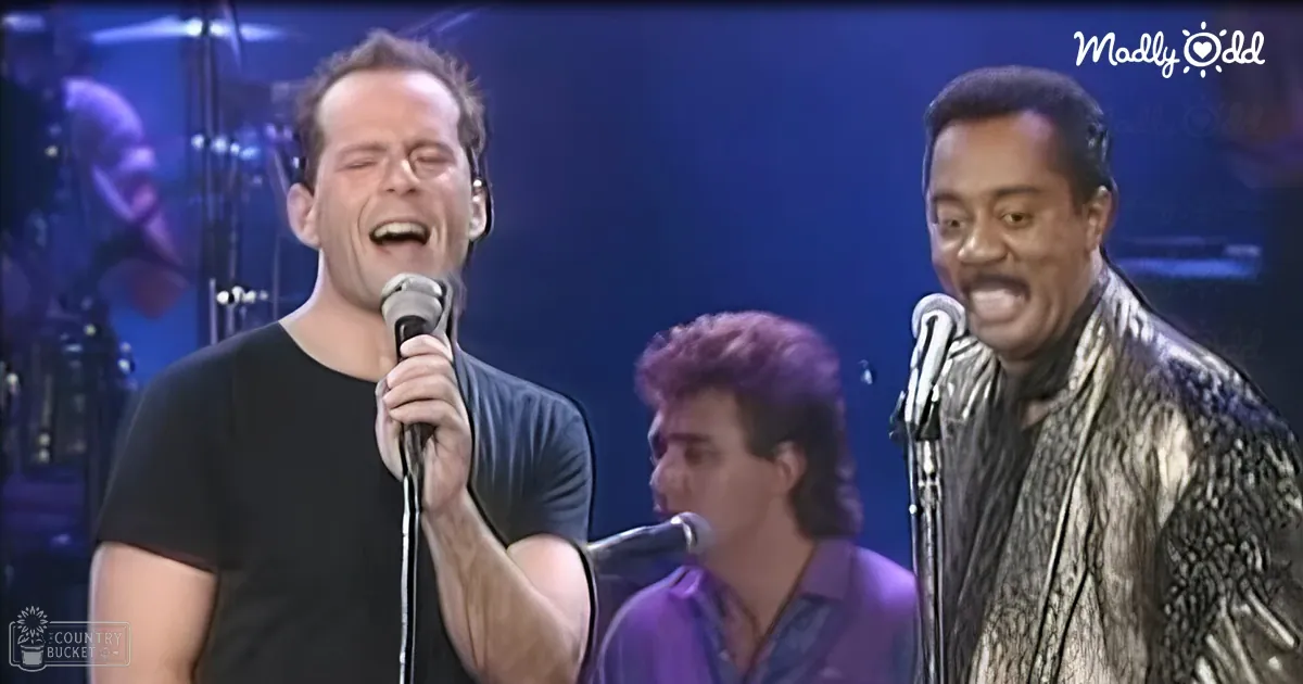 A male singer in a loose-fitting black t-shirt performs on stage alongside another individual holding a microphone; the latter wears a shiny, textured jacket with reflective material.
