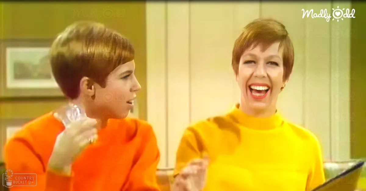 Two children indoors; one child wearing a bright orange long-sleeved top and white gloves, smiling with a clear plastic bottle or container; another adult female in a yellow turtleneck sweater, smiling widely. Setting appears to be a light-colored wooden paneling living room or dining area during the daytime.