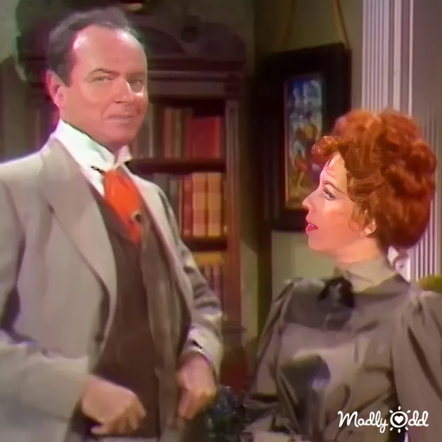 Adult male in a light gray suit jacket, white shirt, and red tie stands indoors with a woman wearing a dark green dress. Bookshelf filled with books is visible behind the man.