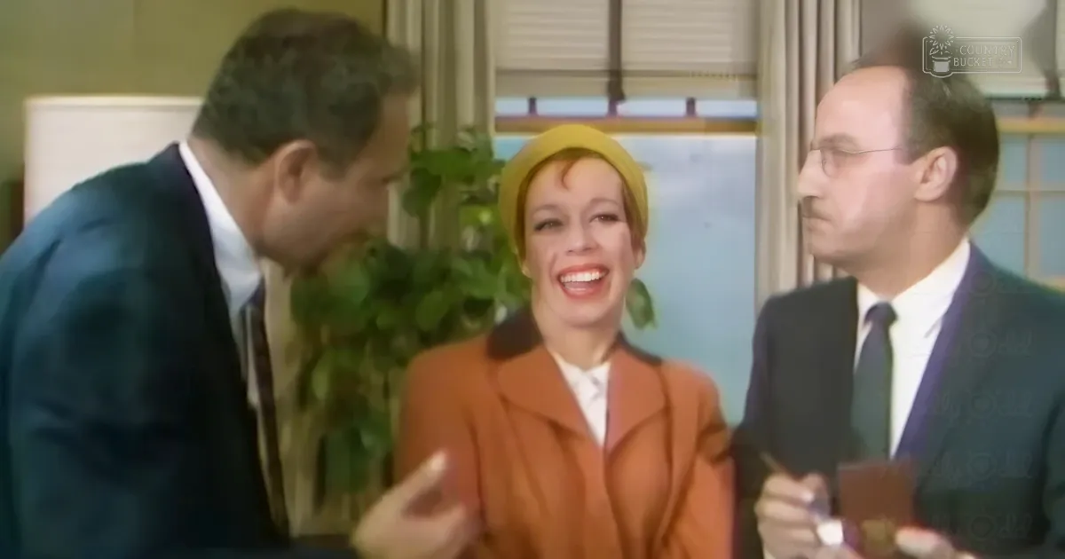 An adult female in her middle years wears a vintage orange coat and yellow hat, seated with an adult male in formal attire in what appears to be an indoor office or professional setting.