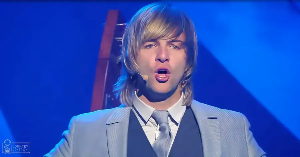 Young adult male in a formal light gray suit performing on a stage with blue lighting at night.