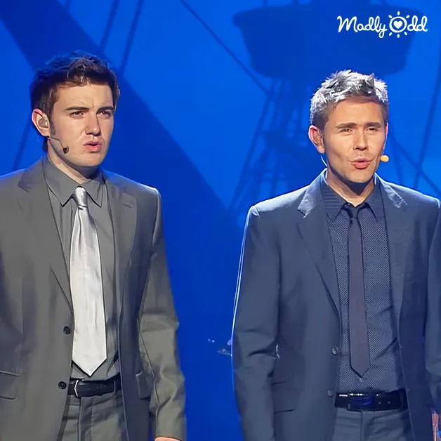 Two young adult men in formal suits stand side-by-side on a stage, wearing well-fitted gray and dark blue suits with light and dark blue ties respectively.