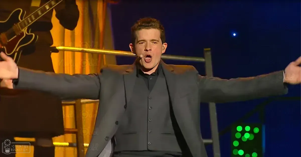 An adult male performer in a dark gray suit jacket and collared shirt stands on stage, surrounded by microphone stand, guitar, decorative lighting with green lights, and yellowish-orange curtains. The setting suggests an indoor performance space like a concert or theatrical show.