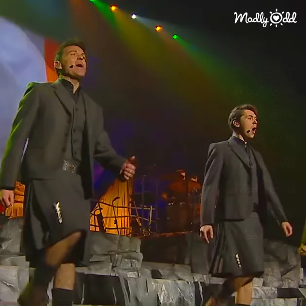 Two performers on a concert or theatrical stage wearing dark-colored knee-length skirts with long-sleeved tops under professional lighting.
