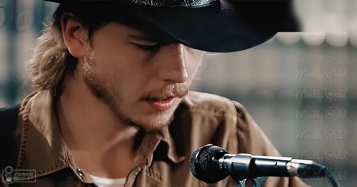A young adult male in a brown jacket with patterned details and black hat sings into a microphone in an indoor evening setting.