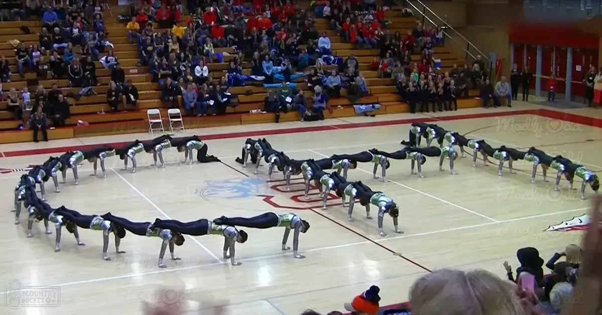 A cheerleading routine in an indoor gymnasium with wooden bleachers and spectators wearing casual attire during fall or winter.