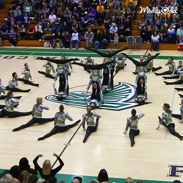 A dance team performs acrobatic moves on a wooden indoor sports arena floor with spectators watching from bleachers.