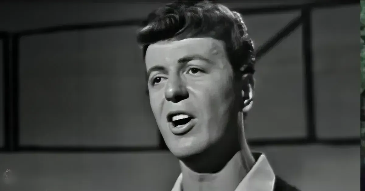 A young adult male wearing a fitted white shirt indoors with artificial lighting.