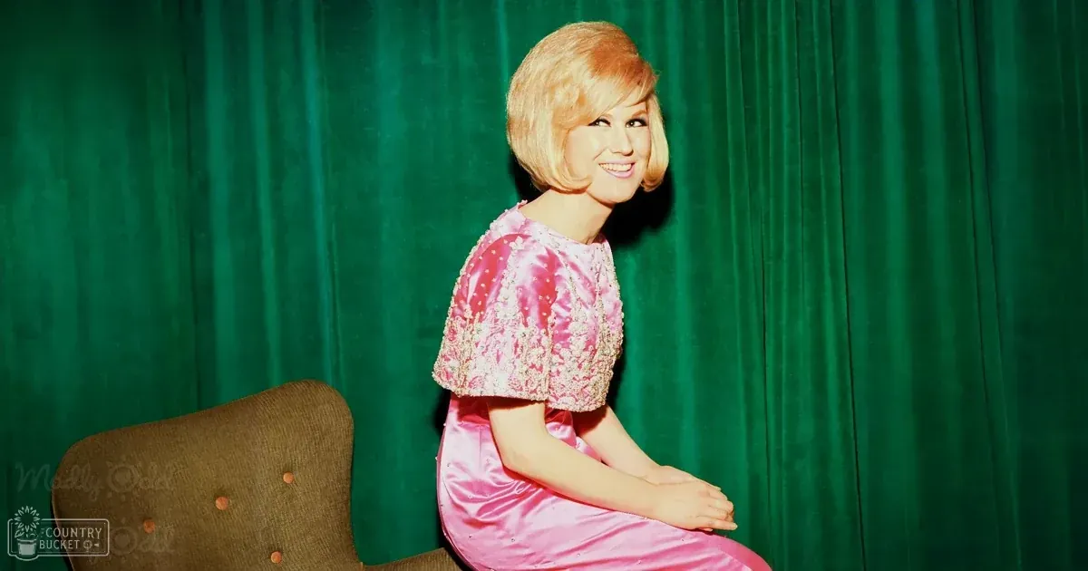 A young adult female in a vintage pink satin dress with floral embellishments, wearing it indoors against green velvet curtains.