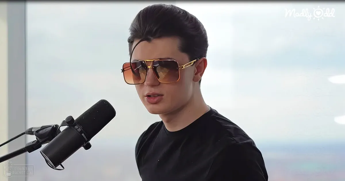 A young adult male wearing a black t-shirt with relaxed fit and round neckline, holding large gold-framed sunglasses, stands indoors near a microphone under an overcast sky.