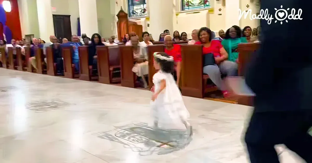 A young girl wearing a white dress with floral headband stands inside a church, surrounded by wooden pews and stained glass windows. Adults in the background are seated on pews dressed casually or semi-formally.