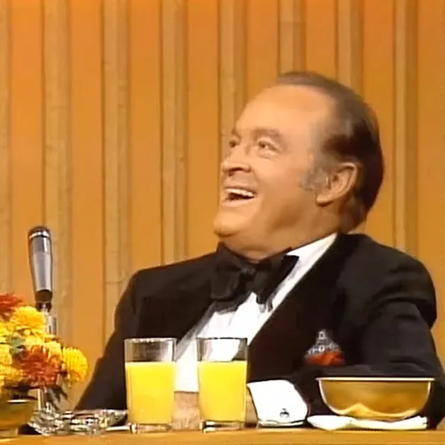 An older adult male wearing a black tuxedo with white shirt and black bow tie, sitting at a table with orange juice glasses and flowers in an indoor studio setting.