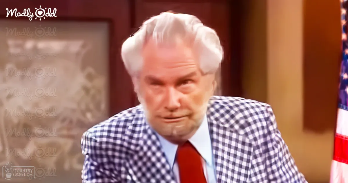 An older adult male wearing a checkered blazer with a white and dark blue pattern, light blue dress shirt, red tie, in an indoor formal setting with an American flag visible on the right.