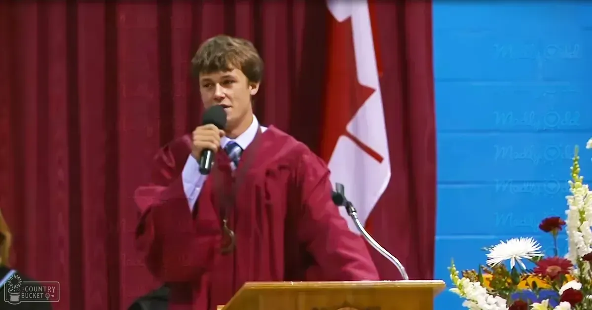 A young adult male wearing a maroon graduation gown with a white shirt and blue tie, holding a microphone at an indoor graduation ceremony in Canada.