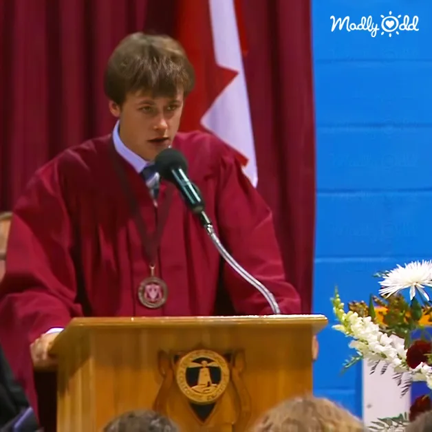 A young adult male giving a speech at a graduation ceremony in Canada, wearing a red gown with white shirt and dark tie, adorned with a medal around his neck. The setting includes a wooden podium with an emblem, floral arrangement of white flowers and greenery, red curtains, and part of a Canadian flag.