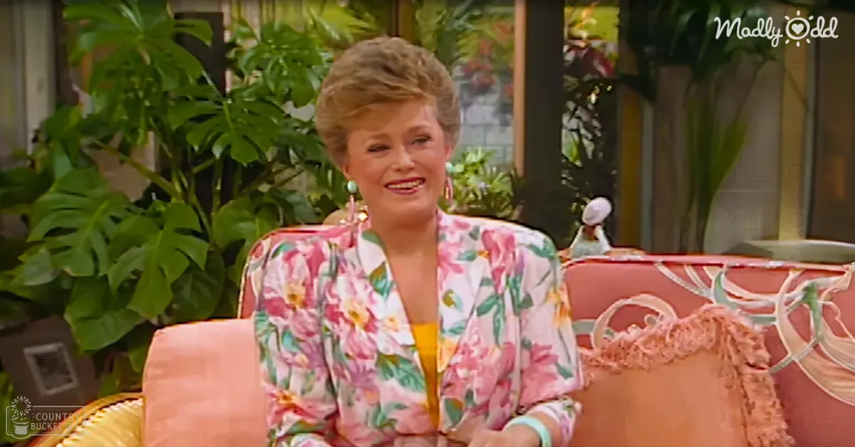 An older adult female wearing a floral-patterned blazer over a yellow top, seated in a room with large windows showing green foliage and pink upholstered furniture.