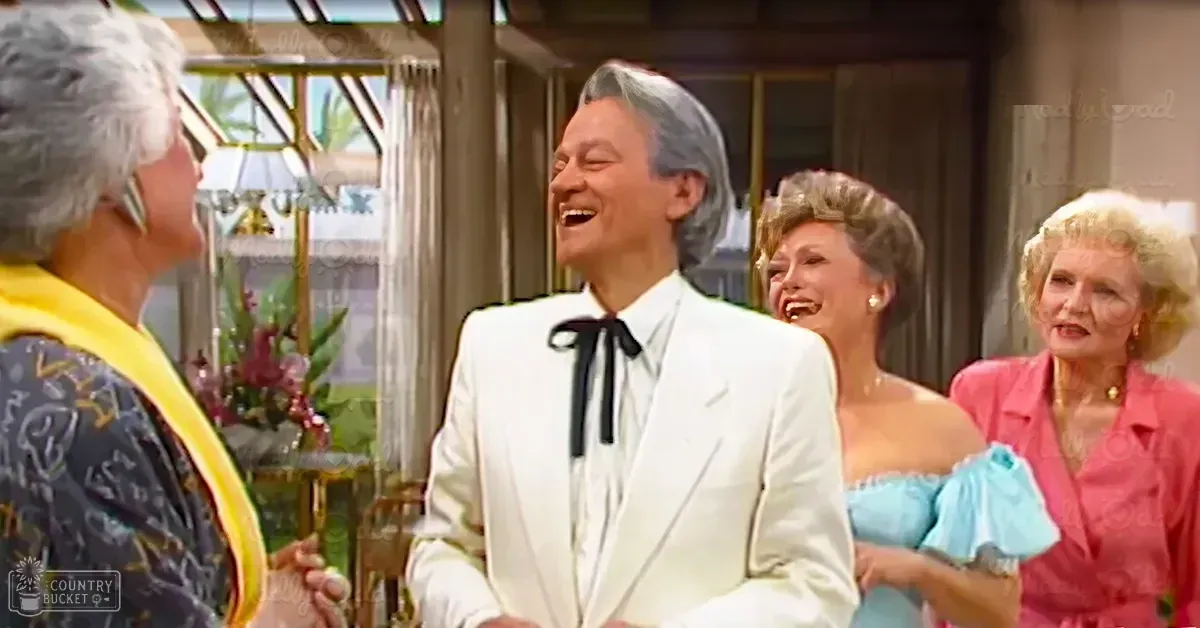 An adult male in a white tuxedo and black bow tie stands indoors amidst large windows with natural light, surrounded by women dressed elegantly in formal attire.