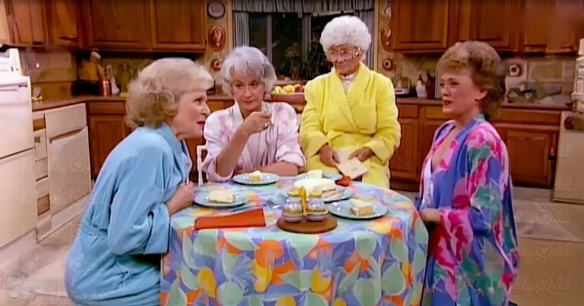Four women in a kitchen setting, wearing light blue, pink floral, yellow, and colorful floral robes with long sleeves, surrounded by wooden cabinets, patterned tablecloth, plates of food, glasses, tea cups, and natural daylight.