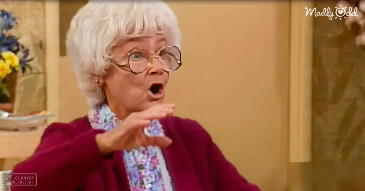 An older adult female wearing a maroon crew-neck sweater, round-framed glasses, and earrings in a well-lit indoor home environment with beige walls and yellow flowers on a wooden shelf.