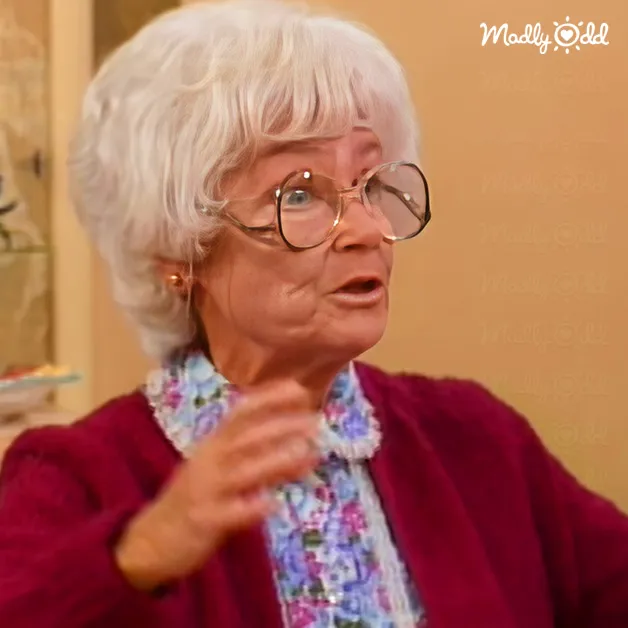 An older adult woman in a domestic setting, wearing a floral blouse with a ruffled collar over a maroon cardigan. The scene suggests daytime indoor lighting.