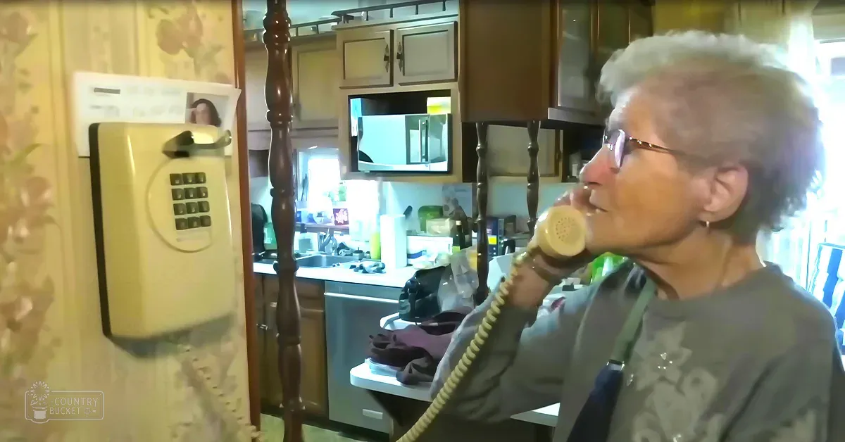An older adult female wearing a dark green long-sleeve shirt in a kitchen setting with beige and brown tones.