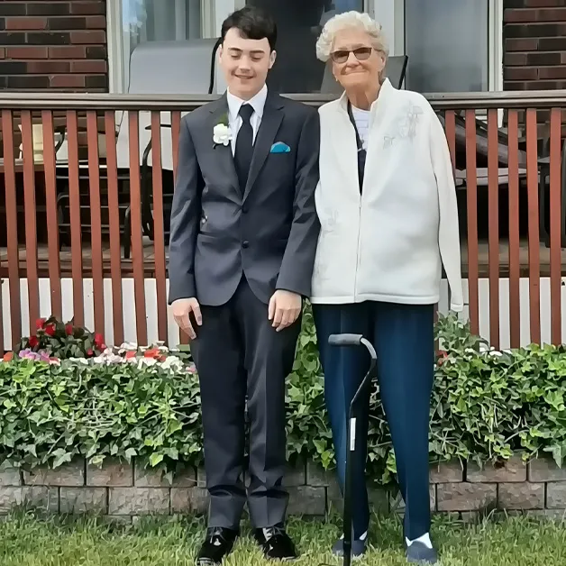 A young adult male in a dark blue suit with a white shirt and black tie stands outside a residential home. An older adult female wearing a white zip-up jacket, dark blue jeans, and black sneakers with white soles uses a black walking cane for support.