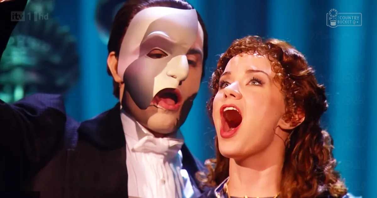 A stage performance from 'The Phantom of the Opera' with two actors, one male in a white mask and black tuxedo, another female in a blue top with gold jewelry. The setting includes teal curtains and greenery, indicative of an indoor theatrical environment.