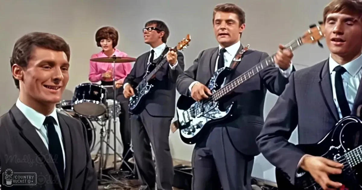 Indoor music performance scene with young adult males dressed formally in dark suits and light blue shirts, set against a plain neutral-colored background.