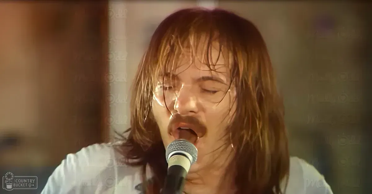 A young adult male sings into a microphone indoors with warm ambient lighting and a cozy setting.