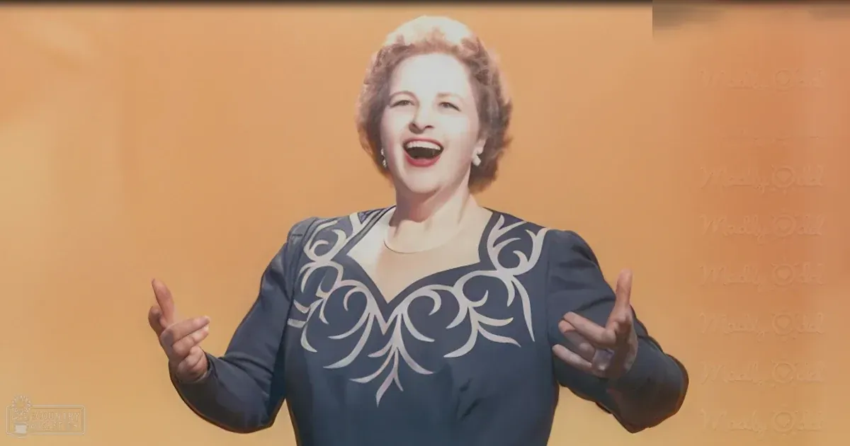 A middle-aged woman wearing a dark blue vintage-style blouse with white floral patterns, pearl-like earrings, and standing against an orange backdrop in what appears to be a professional studio setting.