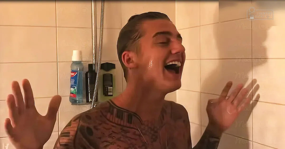 Young adult male wearing patterned shirt with geometric design standing in bathroom setting with light-colored tiled walls and shelves holding toiletries.