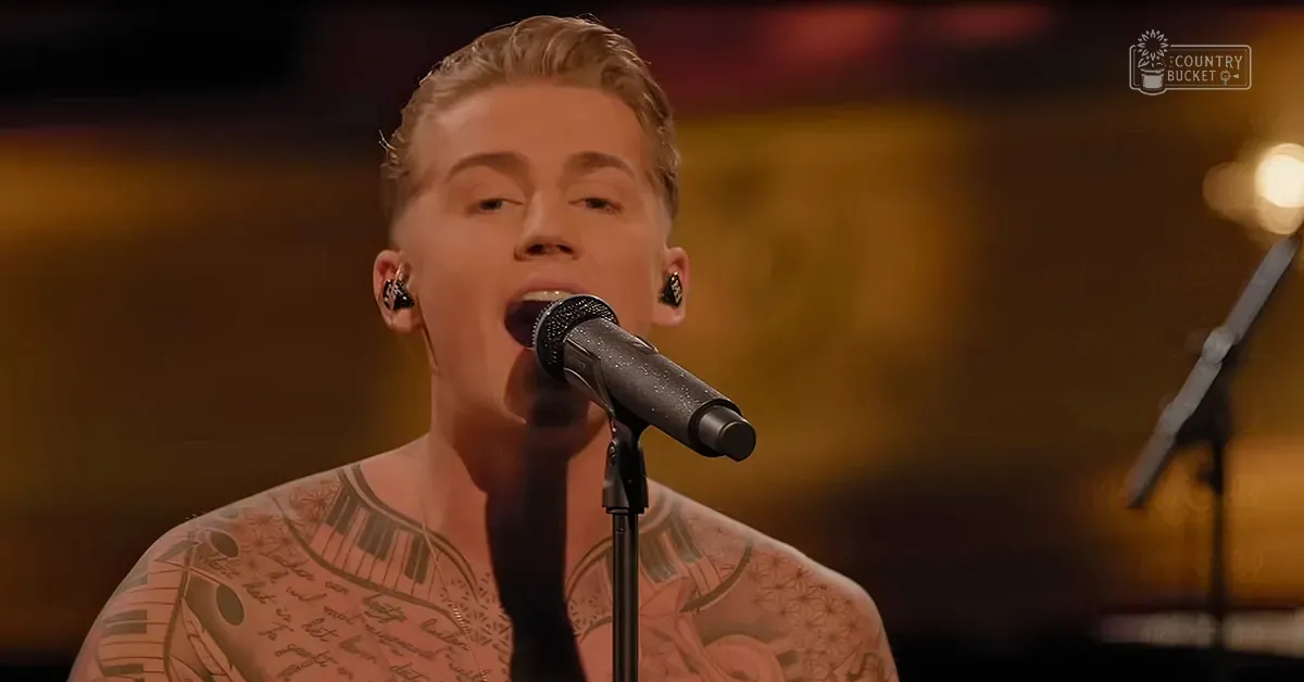 A young adult male wearing a sleeveless top with intricate tattoo designs performs at an indoor venue under warm lighting.