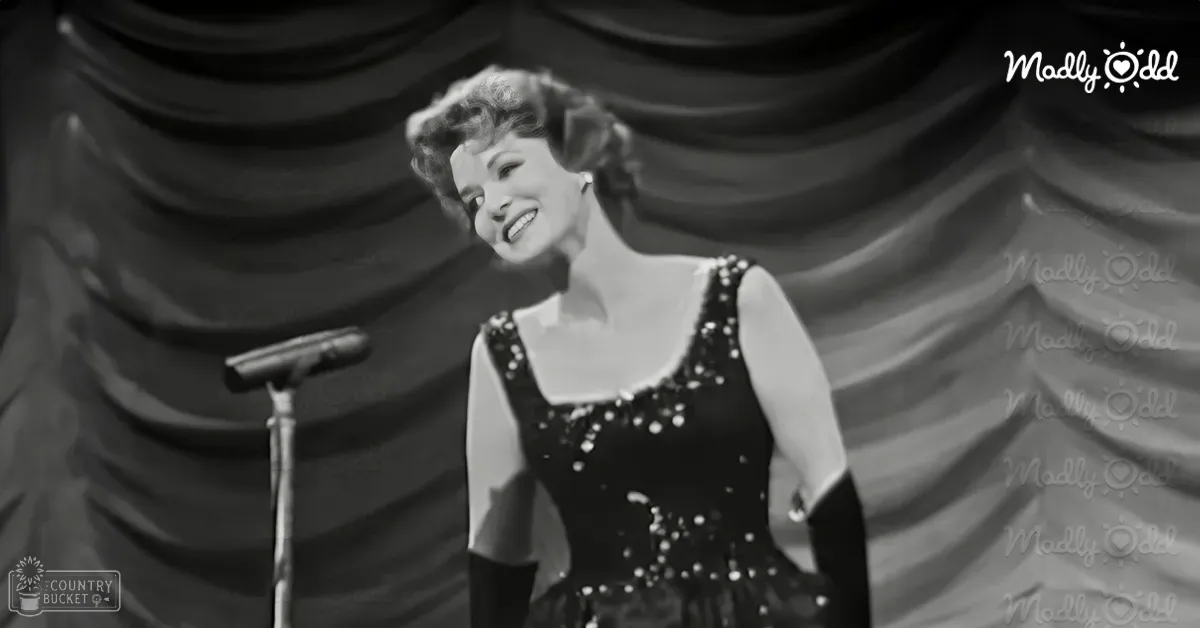 Adult female performer on stage wearing a sleeveless sequined dress, microphone stand visible in front, draped curtain backdrop