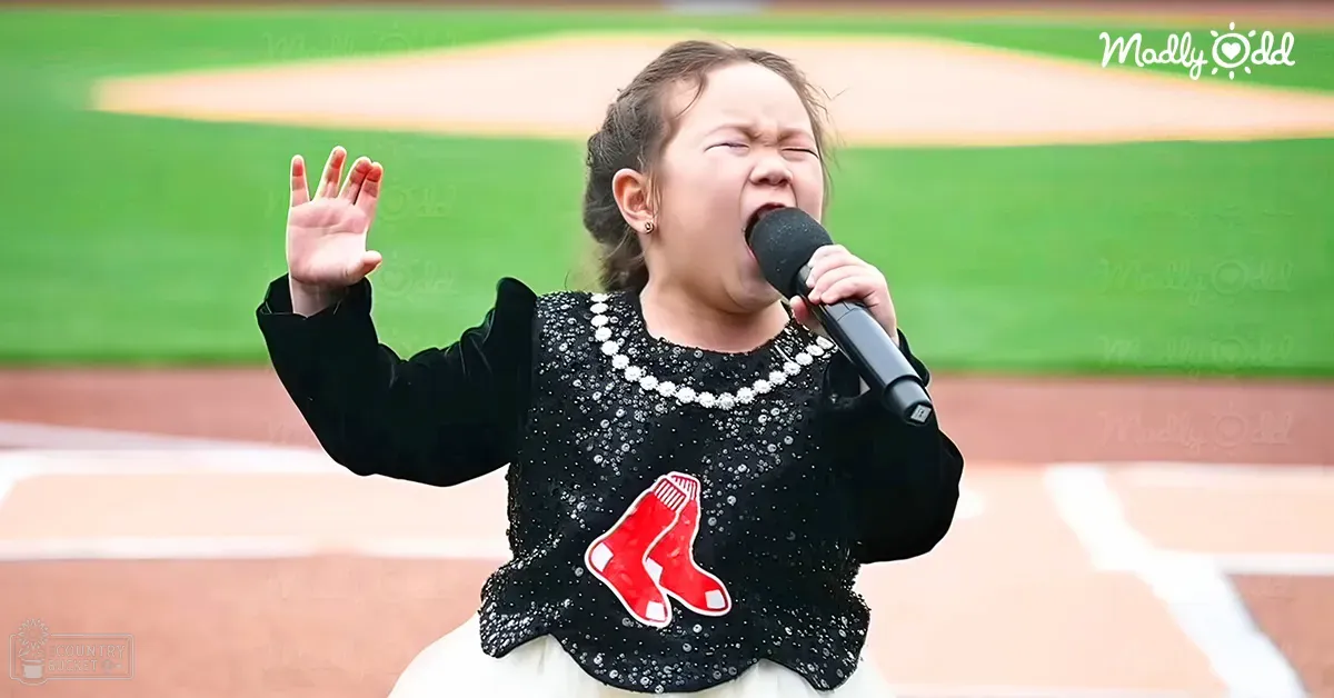 A toddler sings into a microphone at a baseball field, wearing a black, shiny garment with red and white accents.