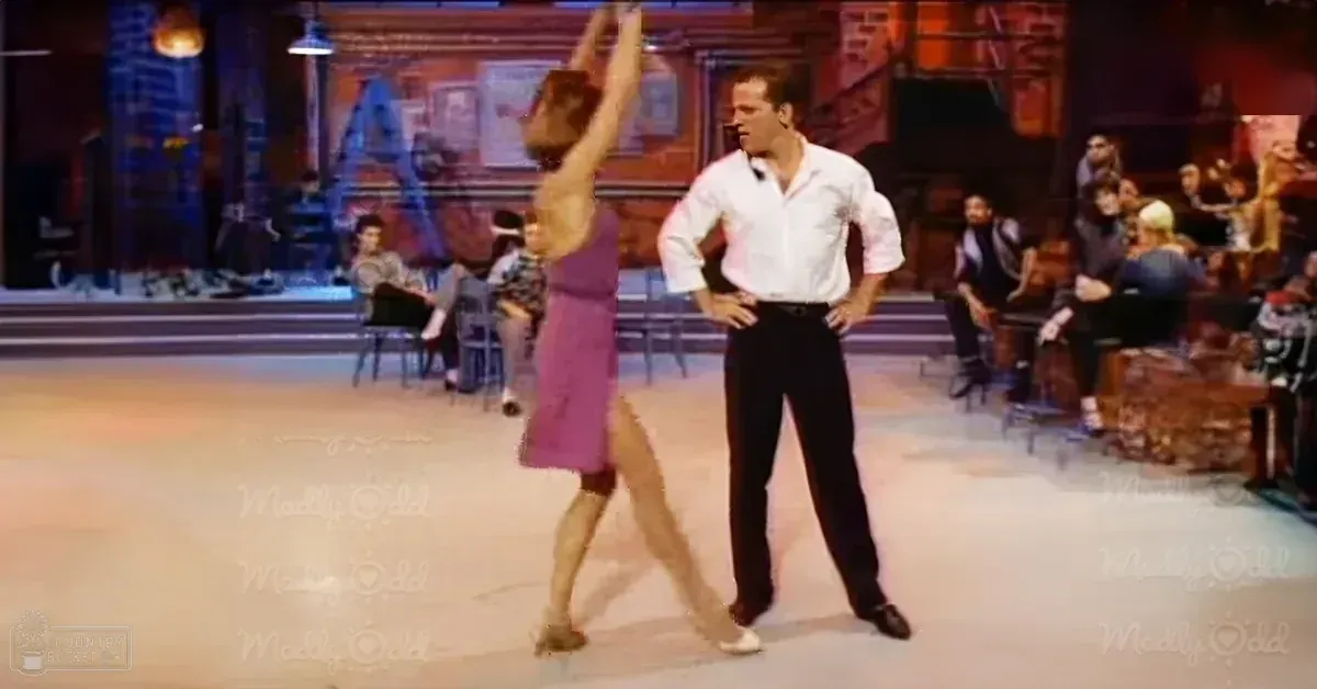 A dance scene with two adult dancers in a studio setting; one male in a white shirt and black pants, the female dancer wearing a purple dress with white shoes.