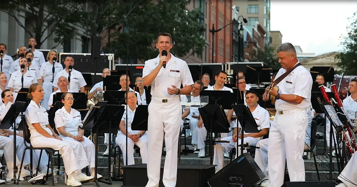 A man in a white naval uniform speaks into a microphone at an outdoor public event with trees and buildings in the background, suggesting spring or summer.