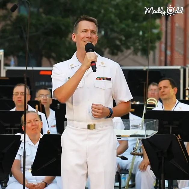 An adult male naval officer speaks into a microphone at an outdoor event with musical instruments and sheet music, set against trees and possibly buildings under soft daylight.