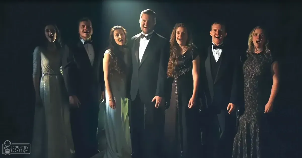 A group of performers in a studio setting, with adults wearing formal attire including white dresses and tuxedos.