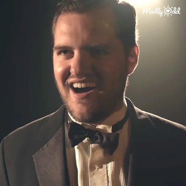 A man in a tailored dark gray suit with a bow tie stands under artificial spotlighting, likely for photography or filming purposes.