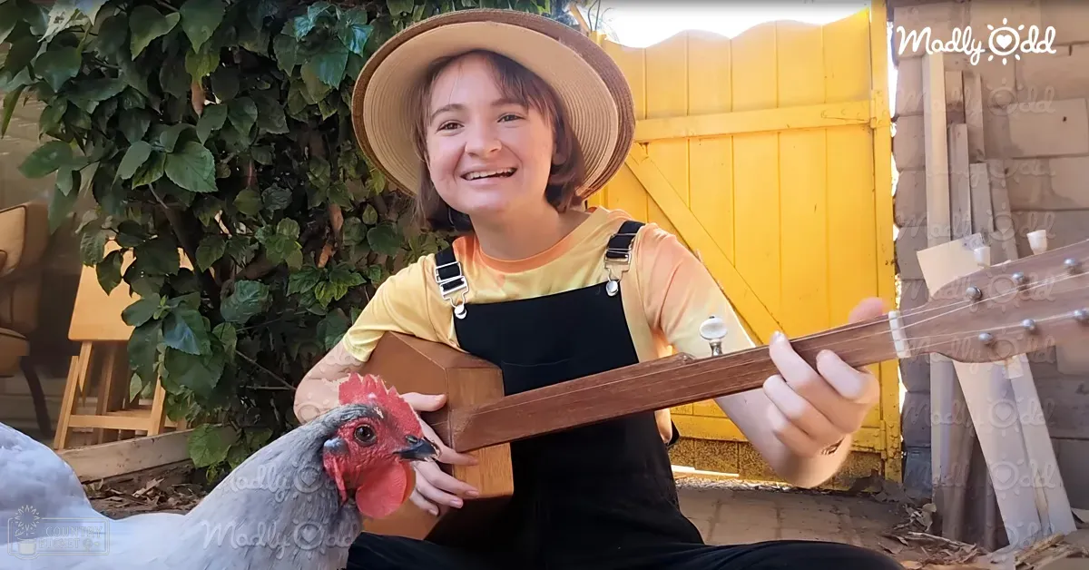 A child in a yellow t-shirt and black overalls with suspenders, wearing a straw hat, stands outdoors amidst greenery. A chicken with gray body and red comb is nearby.