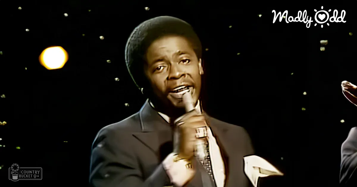 An adult male singer wearing a dark suit jacket and patterned tie from the mid-20th century, singing into a microphone in a studio setting with celebratory decorations.