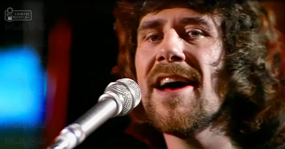 Adult male in casual dark shirt singing into microphone at an indoor venue with blurred stage elements and indistinct decor hints.