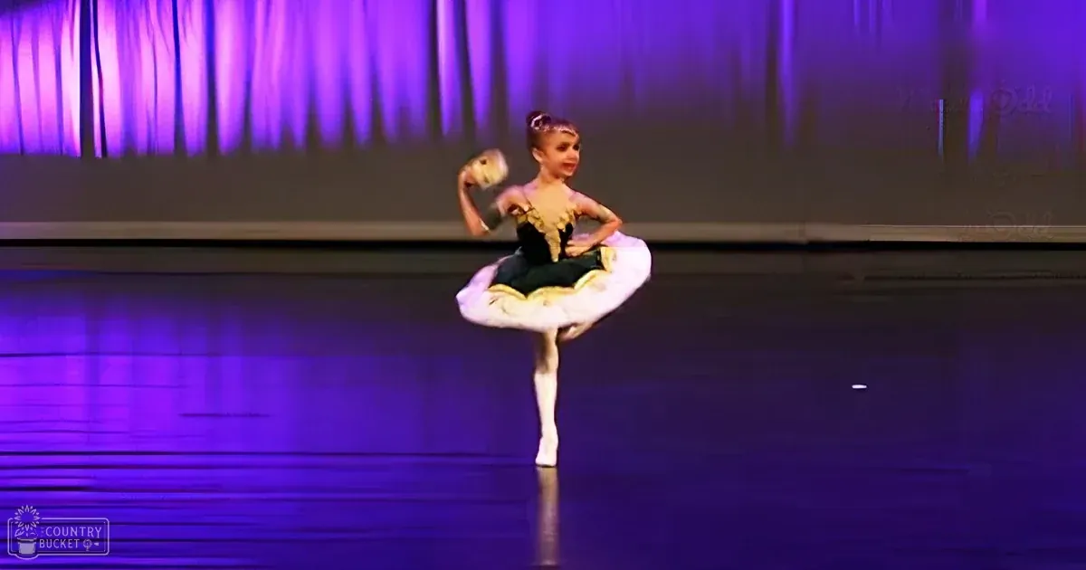 A young child in a black and gold tutu performing on stage with pointe shoes, against a backdrop of purple stage curtains.