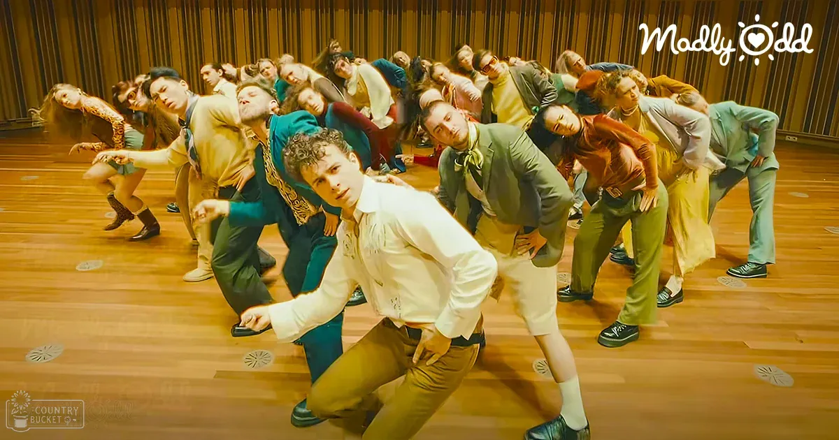 A young adult male stands in an indoor space with wooden flooring, wearing a white long-sleeve shirt with rolled-up sleeves and brown pants, amidst a group of formally dressed individuals.