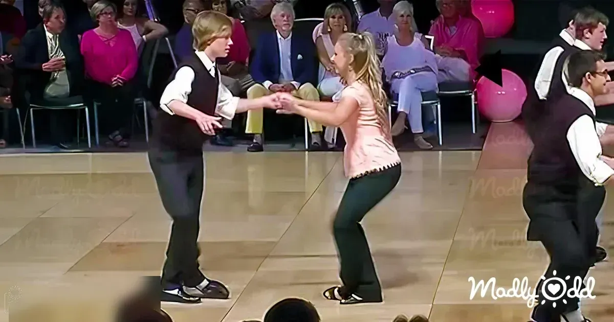 A dance performance with two children in a studio or stage setting with adults as spectators.