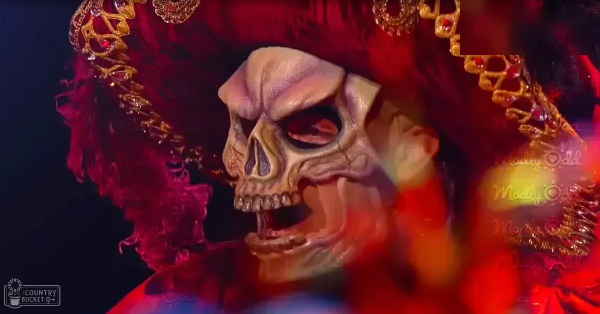 A male performer wearing a costume with a beige skull mask with dark eye sockets and teeth, set against a stage backdrop illuminated by theatrical lighting.