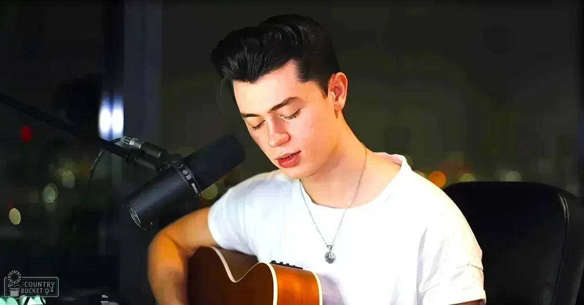 A young adult male wearing a white t-shirt with relaxed fit, modern casual style, and a silver necklace stands indoors against a blurred cityscape backdrop at night.