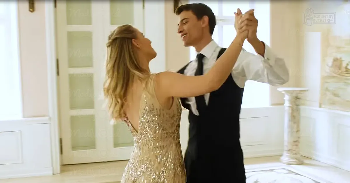 A young adult female wearing a gold sequined dress dances with a young adult male dressed in formal attire, including a black vest and white shirt, in an elegant indoor setting with natural light streaming through frosted glass windows.