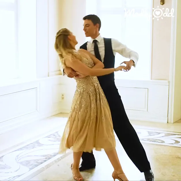 A young adult female in a gold sequined ballroom dress dances with an adult male wearing a black vest and white shirt in a spacious, well-lit indoor ballroom.