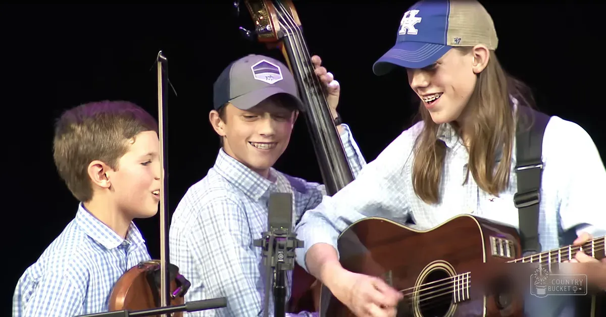 A musical performance scene with children and young adults playing string instruments under focused stage lighting, set against a black backdrop.